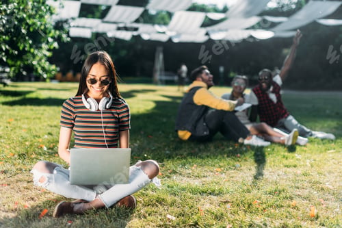 Preview: selective focus of smiling asian woman using laptop and multiracial friends behind resting on green