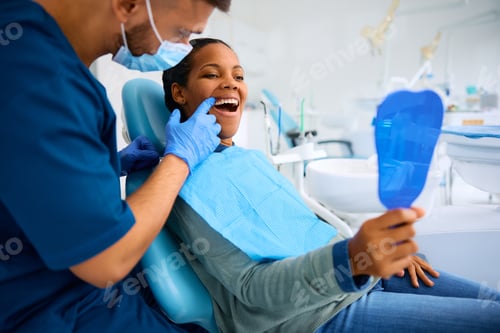 Preview: Black woman using mirror while examining her teeth with a dentist at dental clinic.