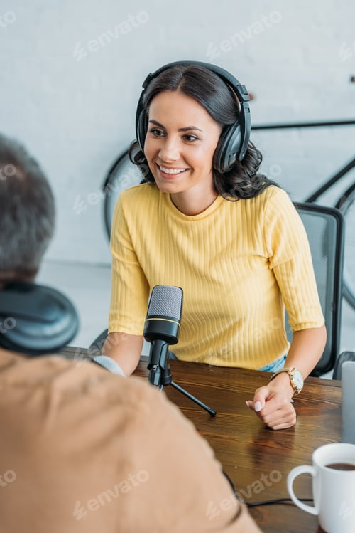 Preview: selective focus of smiling radio host in headphones looking at colleague while sitting near