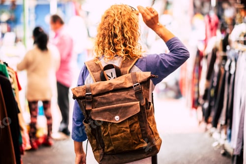 Preview: Blonde curly hair woman backpacker traveler viewed from rear at used market enjoying the shopping