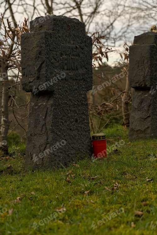 Preview: old cross-shaped tombstone on a graveyard with a red candle of eternal light amidst a green lawn