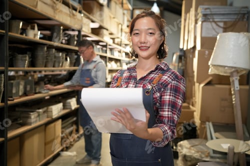 Preview: Smiling woman holding a clipboard in a warehouse with shelves of goods