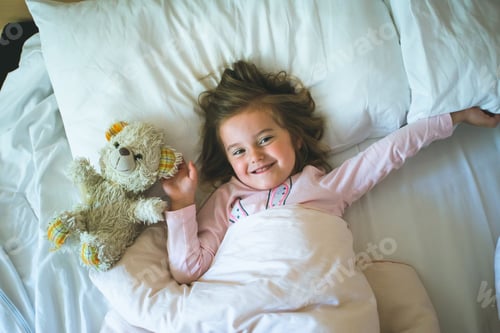 Preview: Little girl lying in a bed with teddy bear in the morning. Having fun at the beginning of the day
