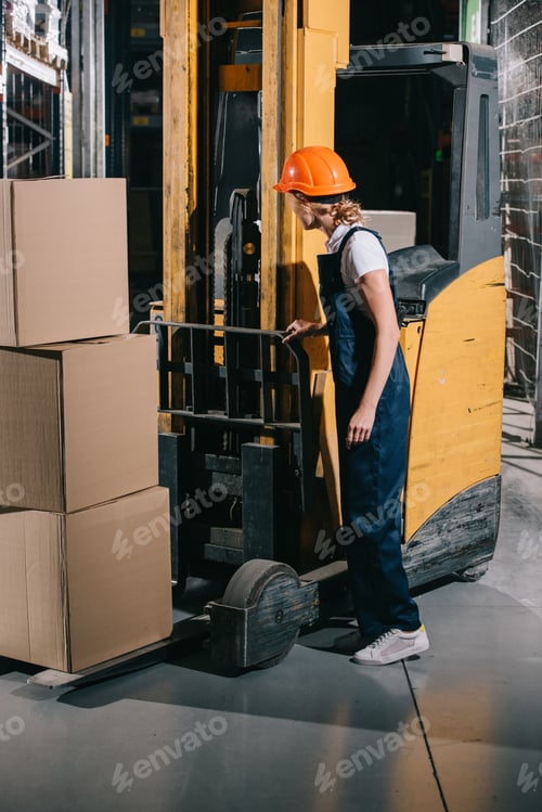 Preview: workwoman in overalls and helmet standing near foklift loader in warehouse