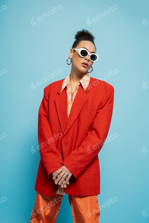 Preview: beautiful african american woman in trendy sunglasses and vibrant outfit posing on blue background