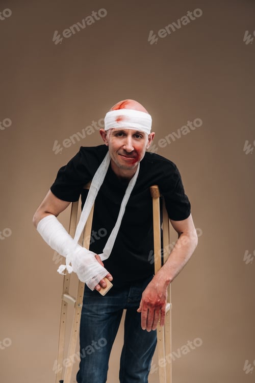 Preview: A battered man with a bandaged head and a cast on his arm stands on crutches on a gray background