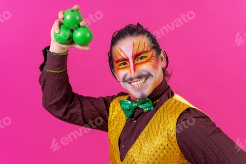 Preview: Clown with white facial makeup on a pink background, juggling green balls