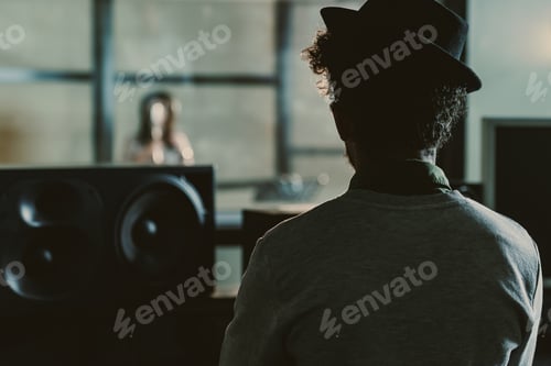 Preview: rear view of sound producer looking at singer recording song behind glass at studio