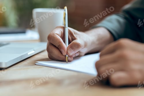 Preview: Hand of mature business man writing some notes in notebook while working with laptop in the office.