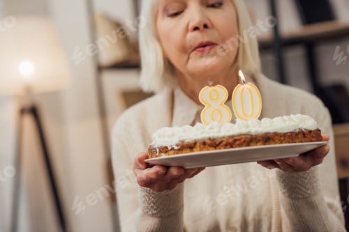 Preview: senior woman blowing out candles on cake with number 80 on top while celebrating birthday at home