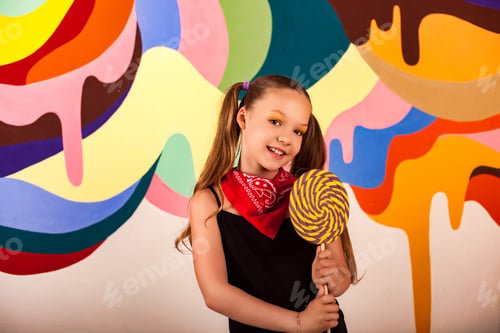 Preview: Portrait of joyful cover child girl with giant round lollipop at colorful wall, happy looking