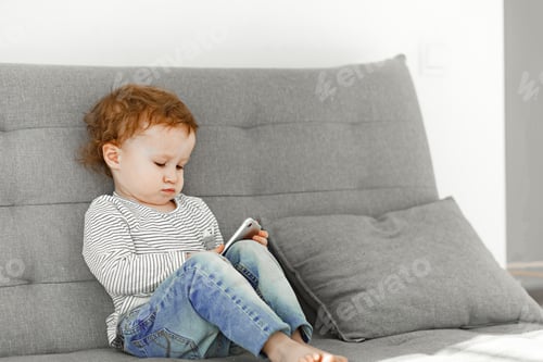 Preview: A little girl in a striped sweater and jeans in a neutral interior holds a phone in her hands.
