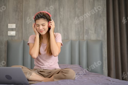 Preview: Asian woman puts on headphones and enjoys listening to music from laptop on the bed in the bedroom