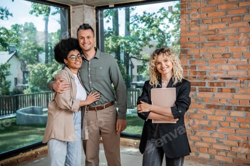 Preview: successful realtor with folder and cheerful multiethnic couple looking at camera in new cottage