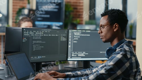 Preview: Sofware developer thinking while touching beard while typing on laptop sitting at desk with multiple
