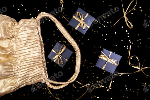 Preview: Black gift boxes with gold ribbon pop out from golden bag on shine background. Flat lay.