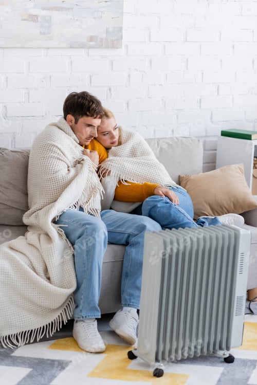 Preview: young couple covered in blanket sitting on sofa and warming near modern radiator heater