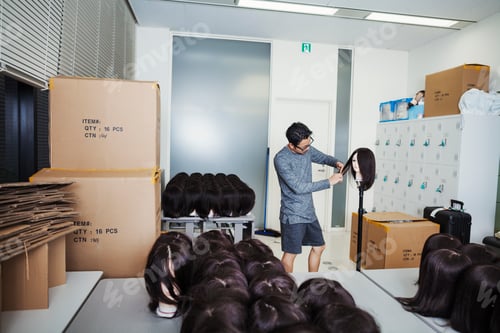 Preview: Bearded man wearing glasses standing indoors, cutting hair of brown wig on mannequin head.