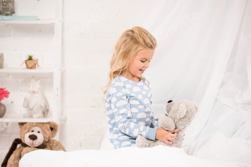 Preview: cheerful kid playing with teddy bear in bed
