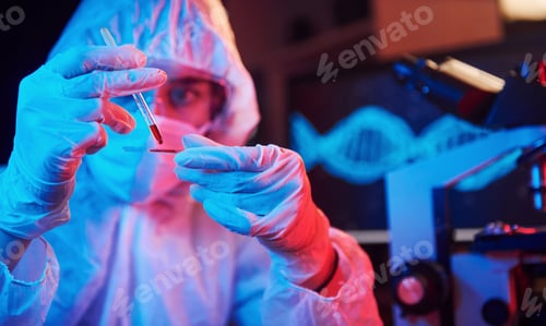 Preview: Nurse in mask and white uniform, holding tube with liquid and sitting in neon lighted laboratory