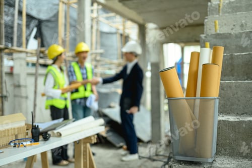 Preview: Rolled architectural blueprints at a construction site with blurred engineers standing in background