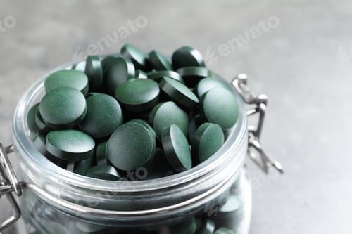 Preview: Glass jar with green spirulina pills on grey table, closeup