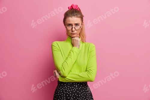 Preview: Sad displeased European woman with pony tail keeps hand under chin and looks unhappily at camera has