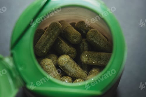 Preview: High angle shot of green capsules inside a bottle on a grey surface