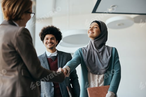 Preview: Happy Muslim businesswoman shaking hands with female colleague in hallway of an office building.