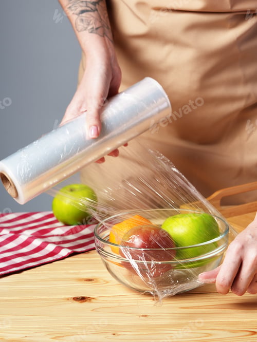 Preview: Woman putting cling filmover bowl of fresh fruit at wooden table
