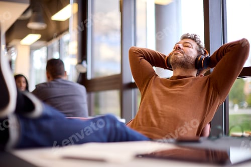 Preview: Mature Businessman With Feet On Desk In Office Listening To Music On Wireless Headphones