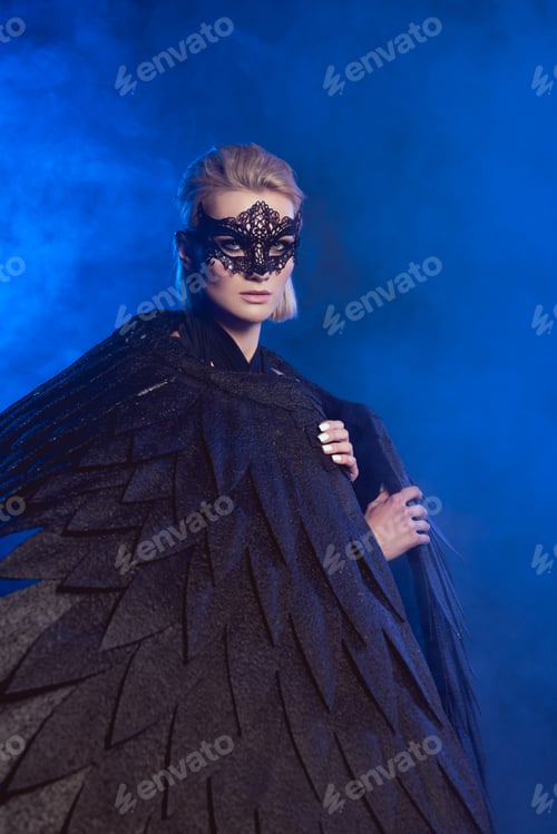 Preview: beautiful mysterious woman with lace mask and black angel wings looking at camera on dark blue