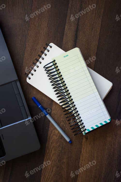 Preview: Top view of a wooden desk with a notebook, and a pen next to a laptop computer.