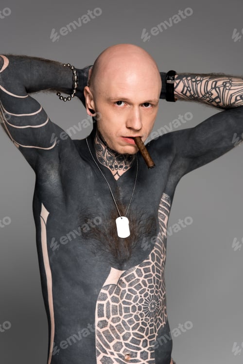 Preview: confident tattooed bare-chested man with hands behind head smoking cigar and looking at camera