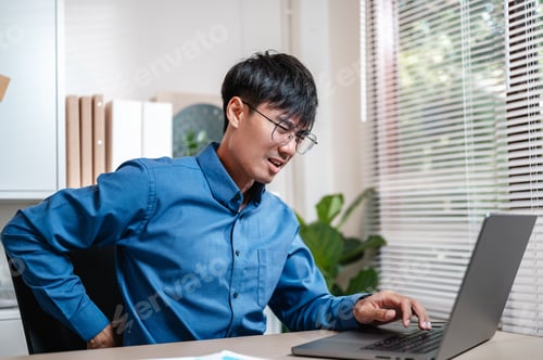Preview: A man in a blue shirt is hunched over a laptop computer. He is wearing glasses and he is in pain