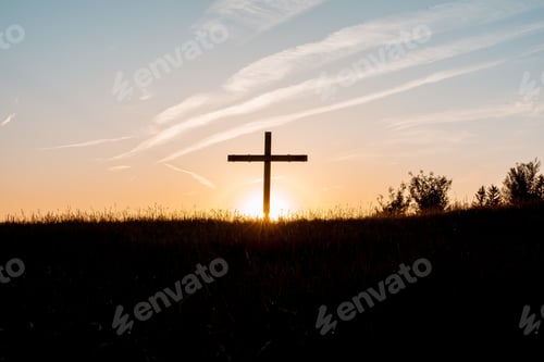 Preview: Silhouette of a wooden cross on a grassy hill with the sun shining in the background