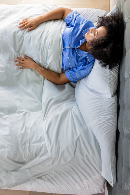 Preview: African American woman lying on bed in bedroom wearing blue pajama top, copy space