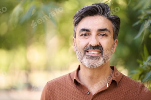 Preview: Relaxed middle-aged man looking at camera while standing outdoors in a park.