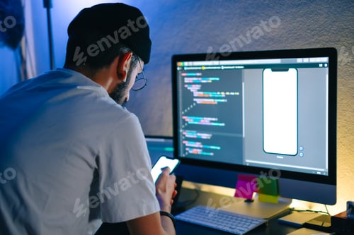 Preview: Young man mobile developer writes program code on a computer and check app in smartphone