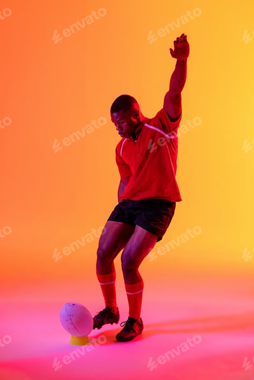 Preview: African american male rugby player kicking rugby ball over pink lighting