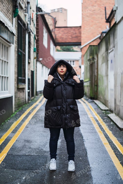 Preview: Vertical shot of a young Caucasian female with a black coat posing in an old neighborhood