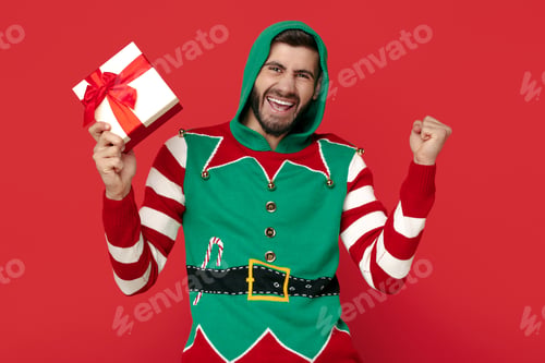 Preview: Happy man dressed in funny Christmas sweater holding gift box and celebrate over red background.