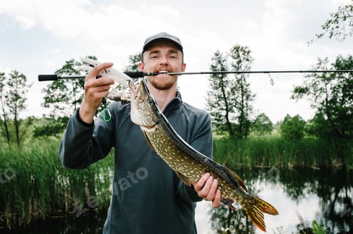 Preview: Happy cheerful young fisherman hold a big fish pike on bridge, pier on a background of lake