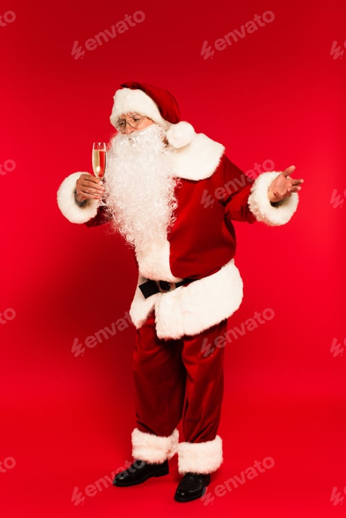 Preview: Full length of santa claus looking at glass of champagne on red background