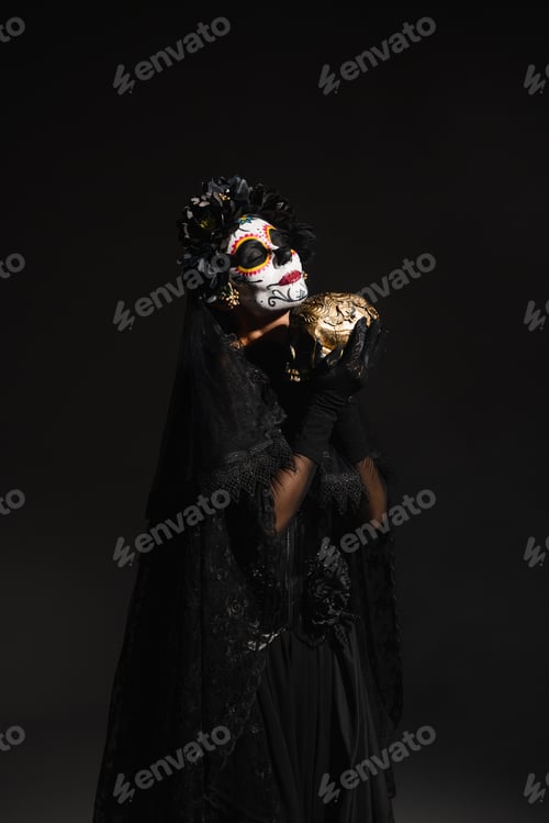 Preview: woman with sugar skull makeup wearing dark costume with veil and holding golden skull isolated