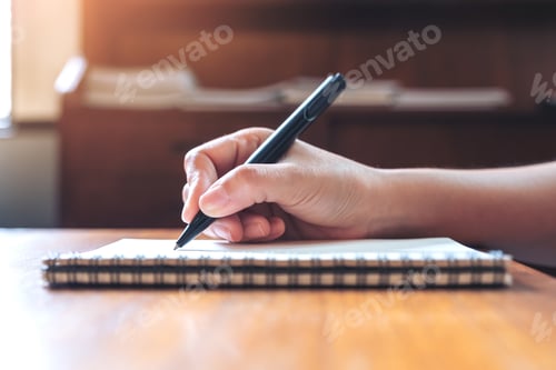 Preview: Closeup image of a hand writing down on a white blank notebook on wooden table