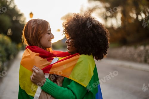 Vista previa: Multiracial lesbian couple look at each other with love embracing with the lgbt rainbow flag
