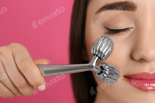 Preview: Woman using metal face roller on pink background, closeup