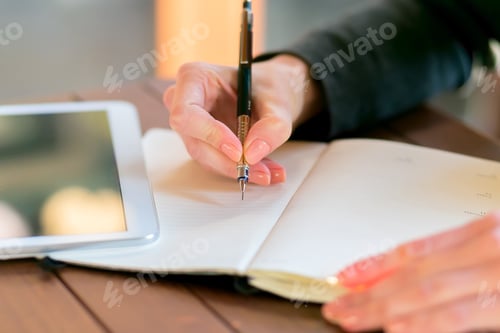 Preview: Person writing notes in a journal at a cafe during daylight hours while using a tablet for reference