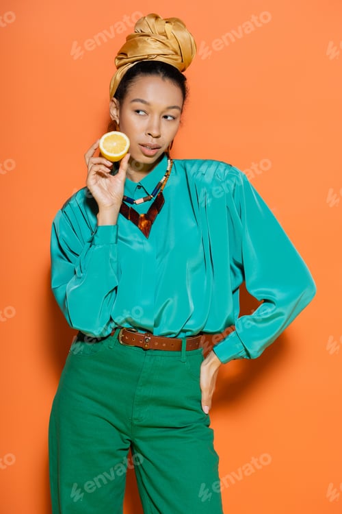 Preview: Trendy african american woman holding lemon and posing on orange background